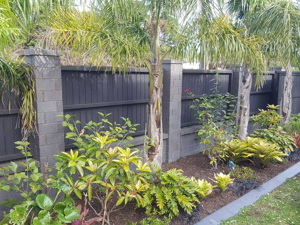 A tidy garden bed of palms, shrubs and mulch along a dark timber fence with block pillars